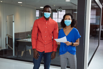 Portrait of diverse male and female colleague in face masks holding paperwork in office