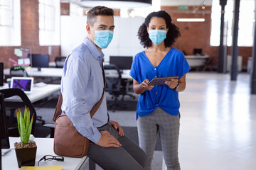 Fototapeta premium Portrait of diverse male and female colleague in face masks standing in office with tablet