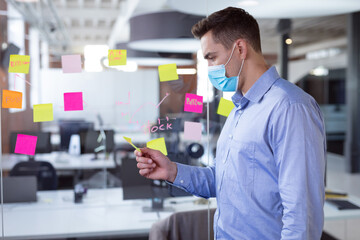 Caucasian businessman wearing face mask brainstorming, reading memo notes on wall standing in office