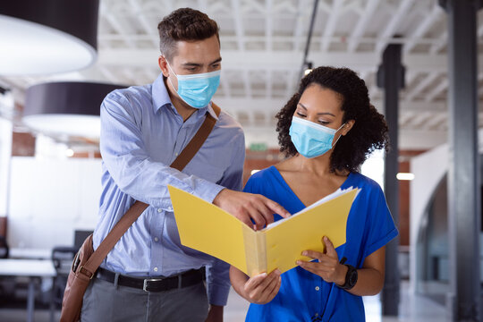 Diverse male and female colleague in face masks discussing paperwork in office corridor