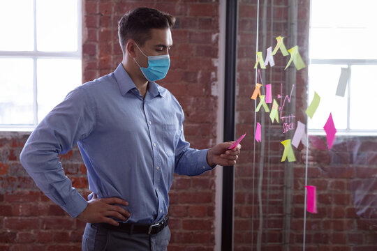 Caucasian Businessman Wearing Face Mask Brainstorming, Reading Memo Notes On Wall Standing In Office