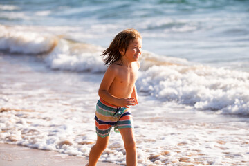 Fototapeta na wymiar Little boy running on beach shore splashing water in blue sea. Kid walking the summer beach.
