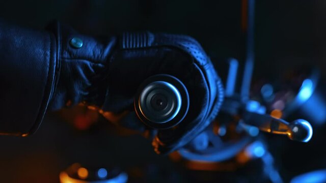 Motorcyclist hand in leather road gloves twists accelerometer lever while standing at city traffic light at night. Multicolored lights illuminate classic motorbike.