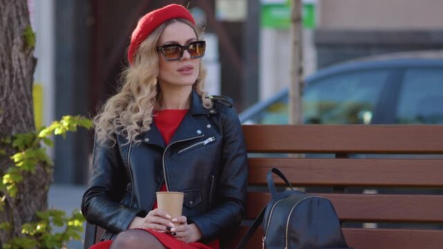 Young beautiful woman in a red beret and dress, black glasses and a braid is sitting on a park bench with cup of coffee. Lunch break at work in the middle of the working day