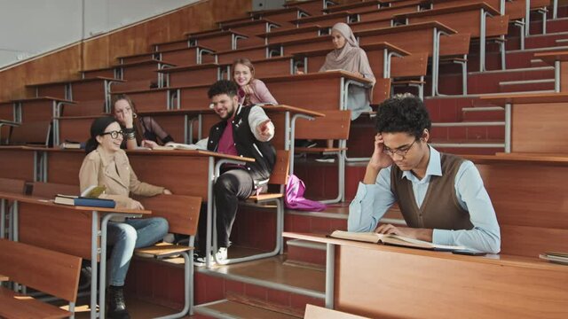 Medium slowmo of group of high school or university students sitting by desks in big auditorium bullying male Mixed-race classmates in eyeglasses pointing at him and laughing while boy reading book