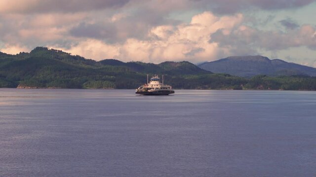 Kanestraum-Halsa ferry crossing. A large ferry carrying cars and passengers across the narrow fjord. The waters are calm. Thick white, pink, and blue clouds, colored by the setting sun cover the sky.