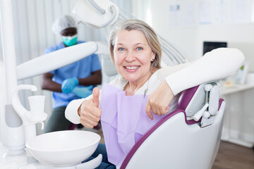 Happy cute mature woman sitting in dental chair after teeth cure giving thumb up. High quality photo