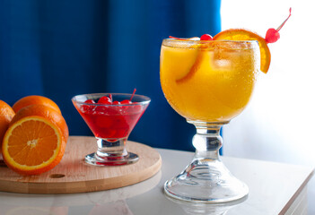 Orange Juicy on a white shiny table with oranges and cherries on the side over a round wooden table