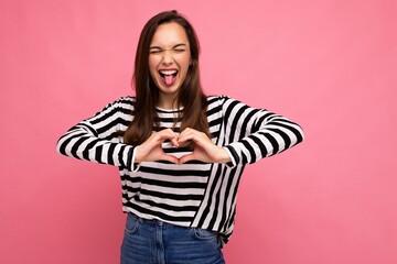 Young positive adorable happy beautiful brunette woman with sincere emotions wearing casual striped pullover isolated on pink background with copy space and showing heart shape with hands. Love