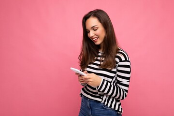 Photo of pretty young smiling brunette woman using mobile phone communicating via texting sms wearing sweater isolated on wall background looking at screen