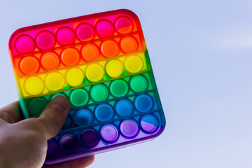 Anti stress toy pop it on blue sky background holding hand. LGBT flag and rainbow colors. A bright thing for children and adults made of silicone.