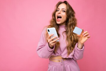 Photo of attractive overjoyed adult blonde curly woman wearing pink clothes isolated over pink background using mobile phone making payment using credit card looking at camera and having fun