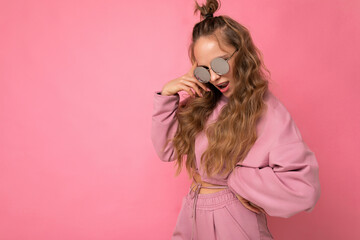 Shot of beautiful happy young dark blonde curly woman isolated over pink background wall wearing casual pink sport clothes and stylish sunglasses looking at camera