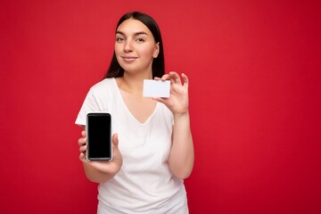 Photo of attractive happy young brunette woman wearing casual white t-shirt isolated over red background with empty space holding in hand mobile phone and showing smartphone with empty screen for