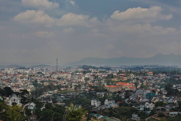 Panorama of the alpine city of Dalat. Many city buildings and green trees. Mountain range against a blue sky with clouds. Vietnam