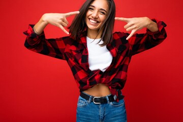 Photo of young happy positive smiling cool attractive brunette woman with sincere emotions wearing white t-shirt and hipster red check shirt isolated on red background with empty space and showing