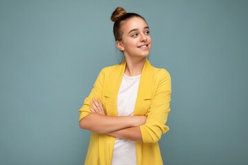 Photo shot of pretty positive happy smiling brunette little girl wearing stylish yellow jacket and white t-shirt standing isolated over blue background wall looking to the side