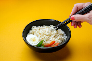 Black bowl of chineese fast cooked noodle with egg and vegetables. Black chopsticks near it at yellow background. Copyspace.