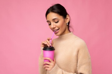 Photo shot of beautiful cute young brunette woman wearing beige sweater isolated over pink background holding paper coffee cup for mockup drinking and looking down