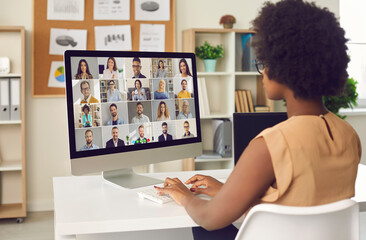 Woman having online business conference or video call staff meeting. Young black lady sitting at office desk with computer talking to team of coworkers who work from home or have hybrid work schedule - obrazy, fototapety, plakaty