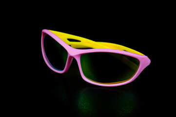 Children's glasses for cycling and sports. Made of pink plastic with yellow temples and UV-resistant lenses. Photographed on a black background.