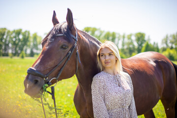 A young rider woman blonde with long hair in a dress posing with brown horse on a field and forest background, Russia