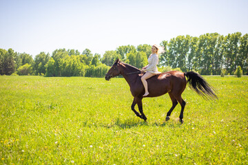 A young rider woman blonde with long hair in a dress riding gallop on brown horse on a field and...