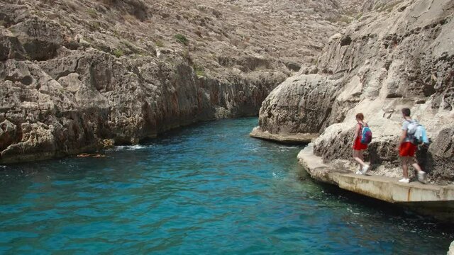 A couple of tourists hiking on the rocky shore of Malta, near the Blue Grotto Cavern.