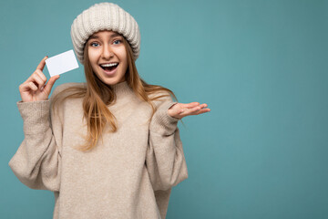 Photo shot of Beautiful amazed positive smiling young dark blonde woman wearing beige sweater and knitted beige hat isolated over blue background holding and showing bank credit card looking at camera