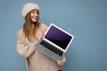 shot of beautiful smiling young dark blond woman in winter warm knitted beige hat holding computer laptop looking at netbook empty free copy space monitor and keyboard wearing beige winter sweater