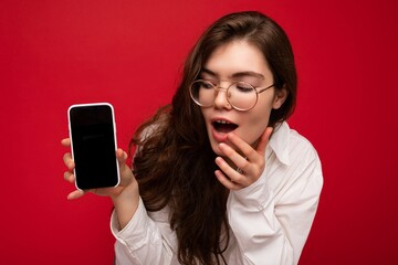 photo of sexy amazed beautiful young brunette woman wearing white shirt and optical glasses isolated over red background holding in hand and showing mobile phone with empty screen for cutout looking