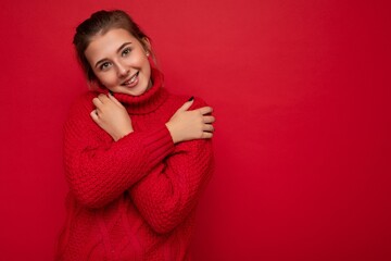 Attractive cute smiling young woman wearing warm red sweater isolated over red background wall keeping hands on chest looking at camera