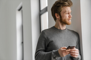 Ginger young man using mobile phone while leaning on wall indoors