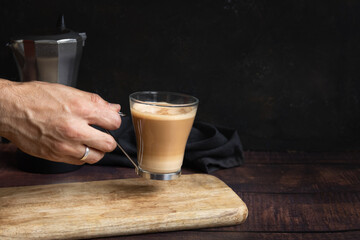 Man's hand holding cup of coffee with milk on wooden table and Italian coffee pot in the background