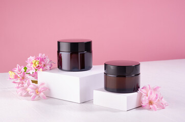 Cosmetic jars for cream of amber glass on white podium and pink wall with fresh spring flowers....