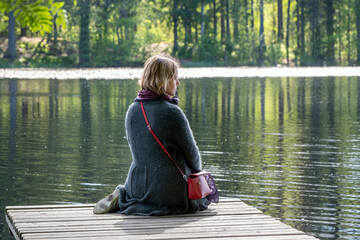 A young woman sits on the wooden pier on a forest lake on a sunny summer day.