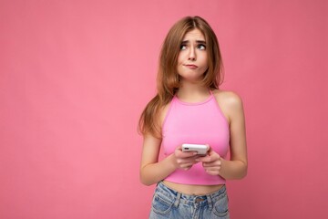 Beautiful thoughtful young blonde woman wearing pink top poising isolated on pink background with empty space holding in hand and using mobile phone surfing on the internet online looking to the side