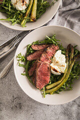 Gerösteter grüner Spargel mit medium rare Rinder Steak Streifen, Rucola, Büffel Mozzarella, Olivenöl und Balsamico in Schale mit Leinen Serviette, Besteck und rustikal Stein Hintergrund