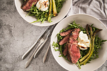 Gerösteter grüner Spargel mit medium rare Rinder Steak Streifen, Rucola, Büffel Mozzarella, Olivenöl und Balsamico in Schale mit Leinen Serviette, Besteck und rustikal Stein Hintergrund