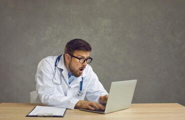 Millennial caucasian male doctor with shocked facial expression sitting at desk looking at laptop...