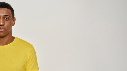 Half face portrait of calm young mixed race man in casual yellow t shirt looking at camera while posing isolated over gray background