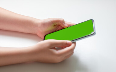 A small child holds a smartphone with a blank green screen in two hands on a white background. Side view, copy space.