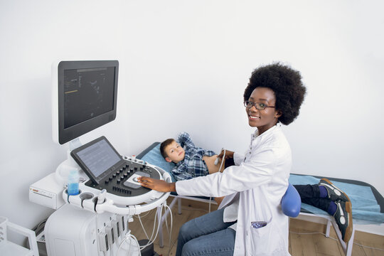 Ultrasound examination of the child in modern clinic. Young likable afro american female confident doctor makes an abdominal scan of a cute school boy in a medical clinic
