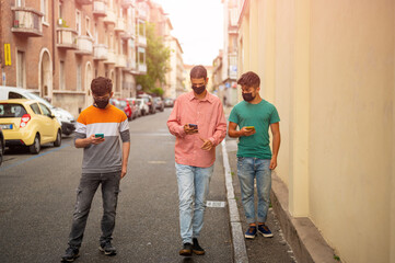 Fototapeta premium Friends group walking on city street with protective black face mask using the smartpjone. Pandemic time. New normal lifestyle concept with young people covered by protective mask. Blurred background.