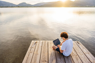 Nerd guy spending time outdoor programming at sunset writing code using laptop. New job opportunity at modern times to work everywhere using notebook and wifi 5g or 4g internet connection technology - obrazy, fototapety, plakaty