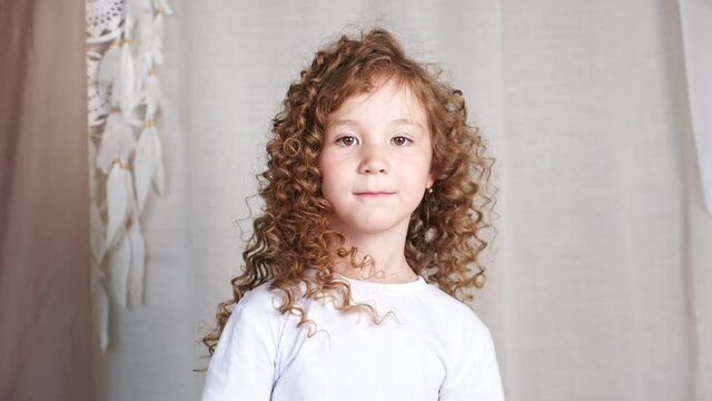 Preschooler little girl with long loose curly hair turns around and smiles slyly looking straight against grey background slow motion closeup