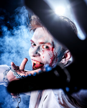 Maniac with makeup and blood on his face, holding a chain with aggression looks into the camera against the background of smoke and glare from the lantern