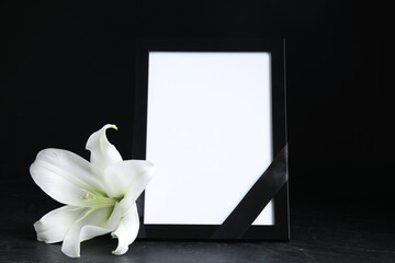 Funeral photo frame with ribbon and white lily on black table against dark background. Space for...