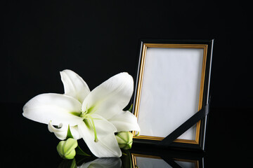 Funeral photo frame with ribbon and white lily on black table against dark background. Space for design