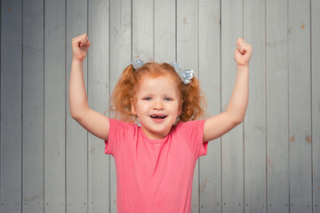 Smiling redhead little girl celebrates success, achieve goal, say yes and raising fists up from joy, triumphing